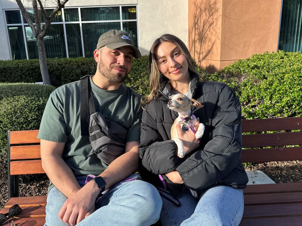 A man and a woman sitting on a park bench with a small dog. The woman holds the dog, while the man wears a green shirt and a cap. They are near shrubs and building windows, reflecting a relaxed atmosphere. This image relates to adoption.