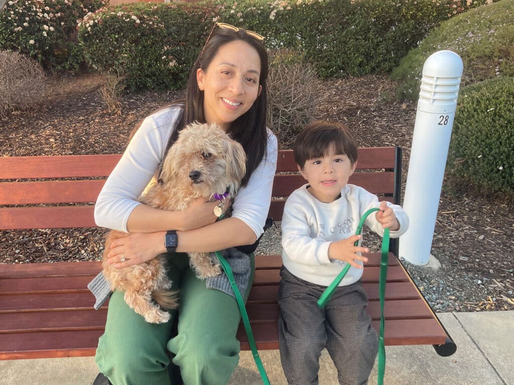 A woman with long brown hair, holding a small dog, sits on a bench beside a young boy. The boy is holding a dog leash and wearing a light grey sweater. They are in a park setting with greenery in the background, related to adoption.
