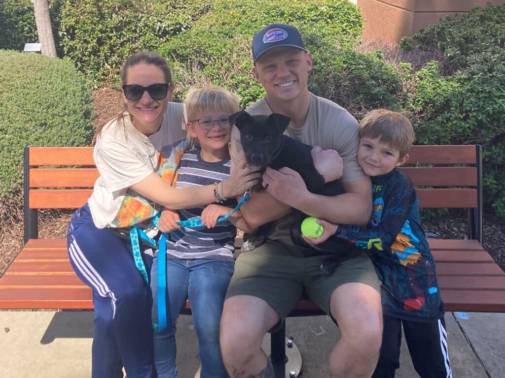 A family of four sitting on a park bench, smiling while embracing a black dog. The mother and father, along with two young children, are dressed casually. The scene reflects a joyful moment, suggesting an adoption celebration.