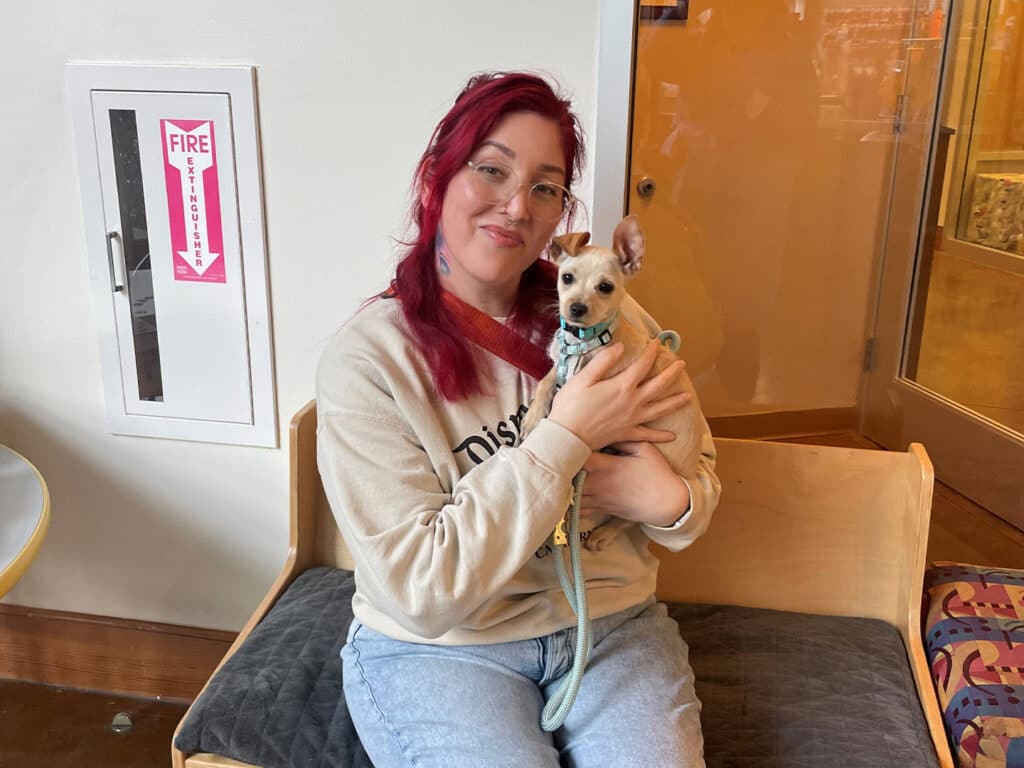 A woman with red hair wearing a beige sweatshirt is seated on a bench, holding a small tan dog. The dog is wearing a leash. The setting features a wall with a fire safety sign nearby.