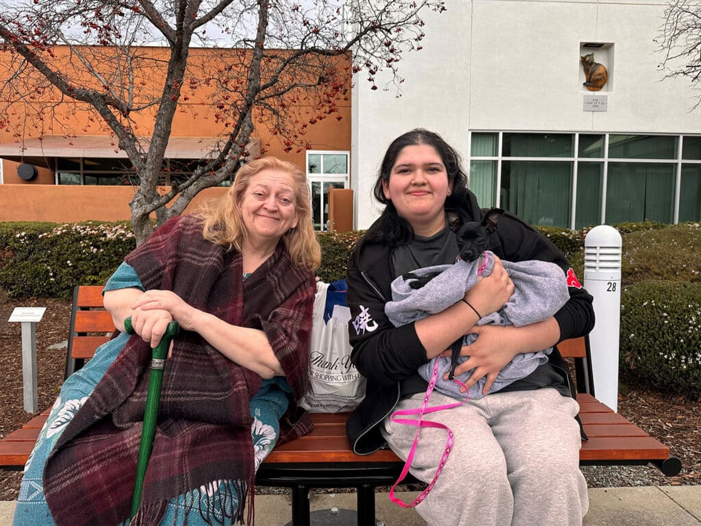 A woman and a girl sit together on a wooden bench outside a building. The woman is holding a cane, wrapped in a plaid blanket, while the girl holds a small animal swaddled in a blanket. Trees with buds surround them, suggesting early spring.