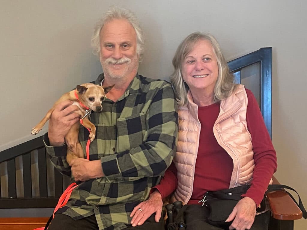 A man and a woman sit together on a bench, both smiling. The man holds a small dog in his arms, and the woman has her hands on her lap. They are dressed casually, surrounded by a simple indoor setting. This image conveys themes of companionship and adoption.
