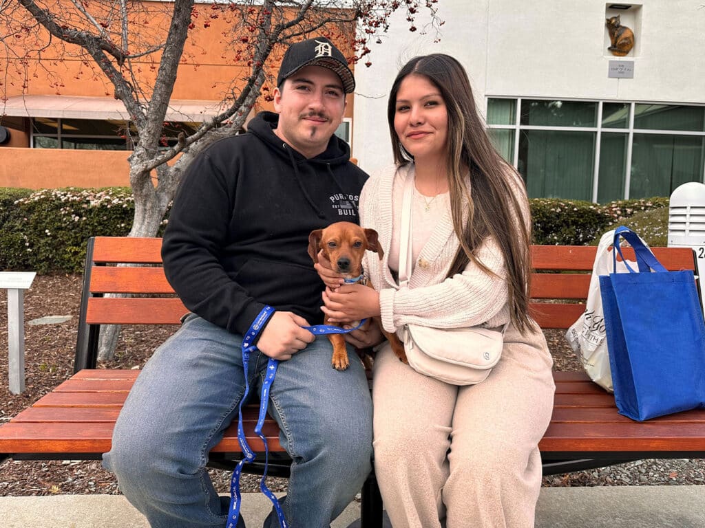 A young man and woman sitting on a bench, holding a small brown dog, in a park-like setting. The woman has long hair and is dressed in a cream-colored outfit, while the man wears a black hoodie and blue jeans. Nearby, there are trees and buildings.