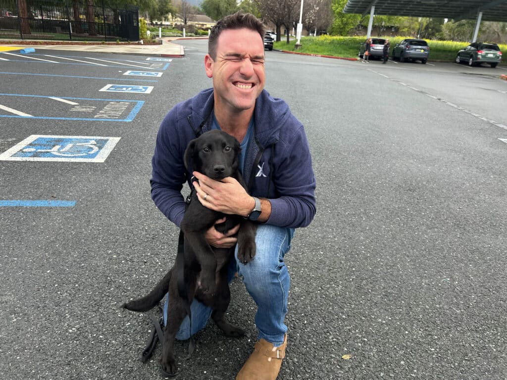 A man kneeling in a parking lot, smiling widely while hugging a black Labrador puppy. The background features parked cars and greenery in the distance, emphasizing an outdoor environment related to pet adoption.