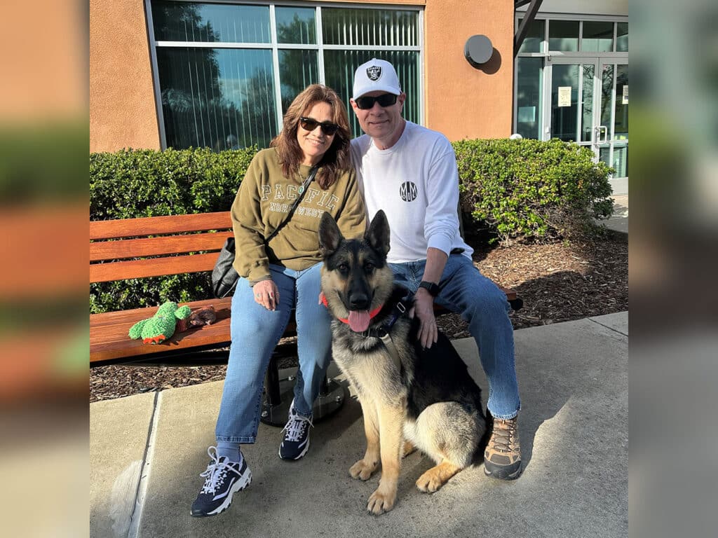 A woman in a green hoodie and a man in a white shirt and cap sit together on a bench outside a building, with a German Shepherd dog in front of them. The scene suggests a moment of companionship, relevant to themes of adoption.