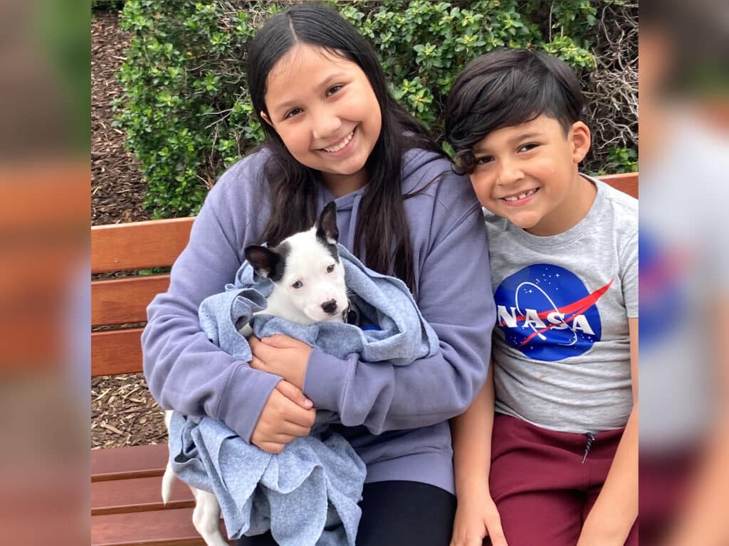 A girl holding a small white puppy wrapped in a blue blanket sits next to a boy wearing a NASA shirt on a bench. They are smiling, with greenery in the background, highlighting a moment related to pet Adoption.