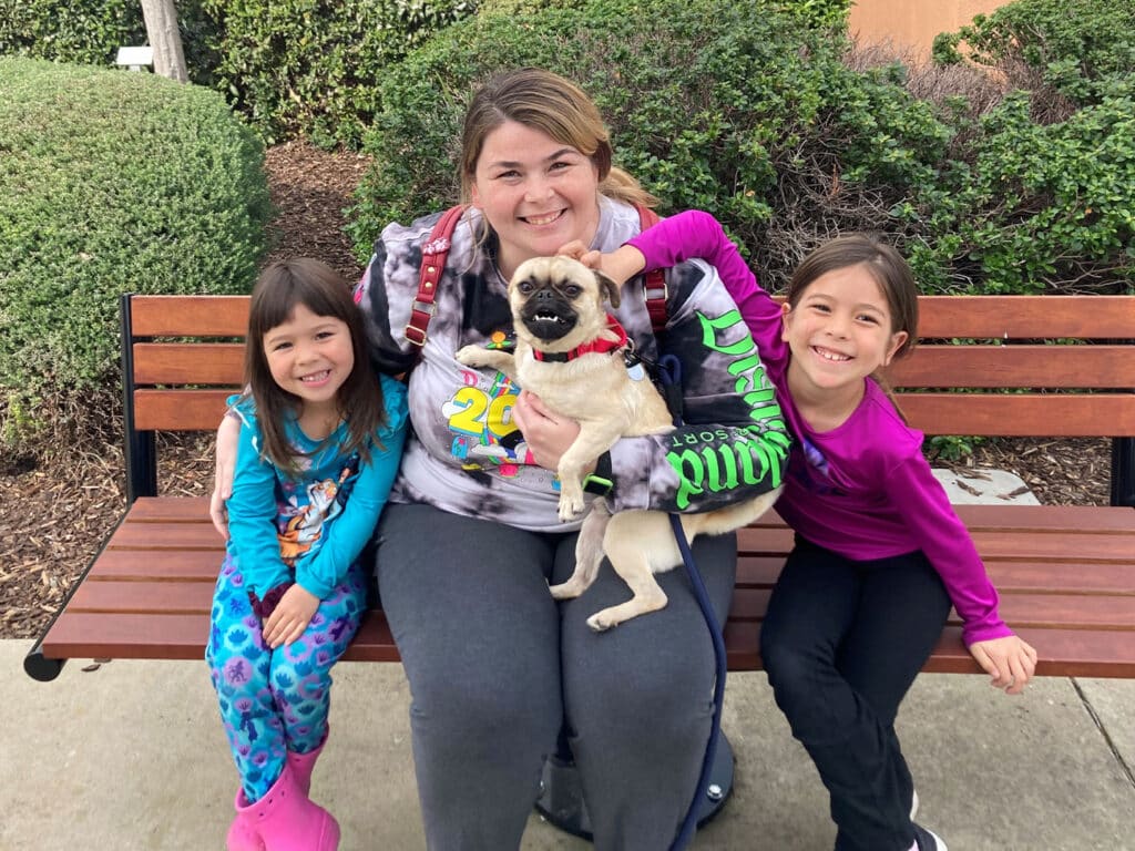A woman sitting on a bench holds a small pug dog while two young girls sit beside her, smiling. The girls wear colorful pajamas. In the background, there are neatly trimmed bushes, contributing to a family-friendly atmosphere related to pet adoption.
