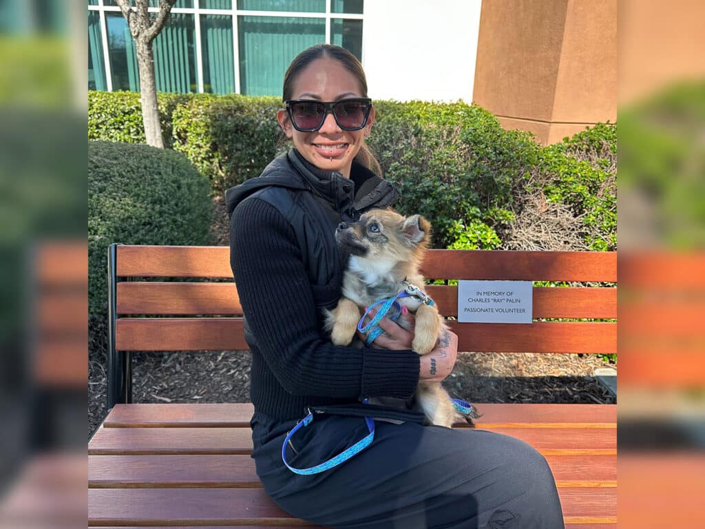 A woman wearing sunglasses sits on a bench, holding a small dog in her lap. The dog looks up at her. In the background, there are bushes and a building. This image emphasizes themes of adoption and companionship.