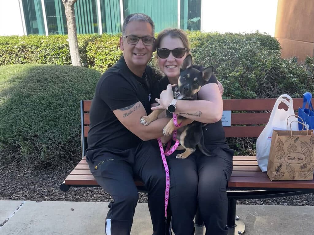 A man and a woman sit on a bench, smiling while holding a small dog. The woman has sunglasses on and is wearing a light shirt. The man is dressed in a dark t-shirt. They appear happy, surrounded by greenery and bags suggesting a recent adoption.