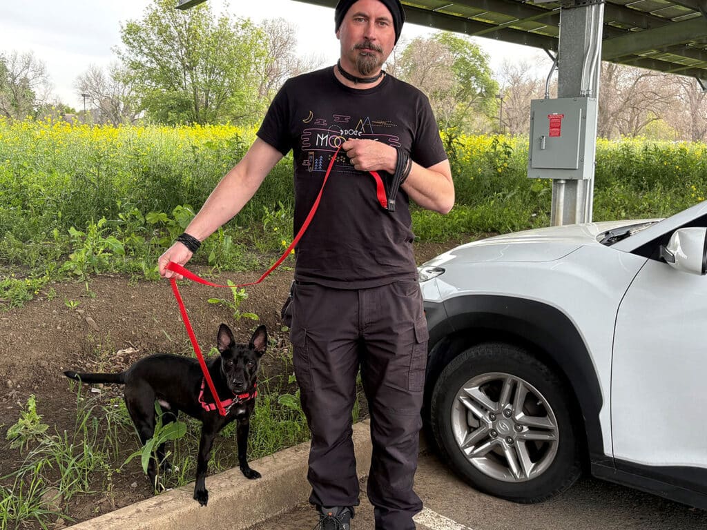 A man standing next to a black dog on a leash at a shaded parking area. The dog is wearing a harness. In the background, there are green plants and trees. This image highlights a moment related to pet adoption.