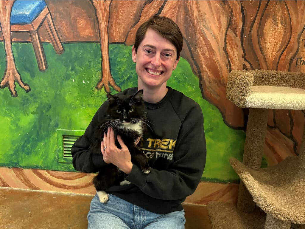 A person smiling while holding a black and white cat in a cozy indoor setting. The background features a mural of trees and a chair, adding to the relaxed atmosphere. This moment highlights the joy of pet adoption.