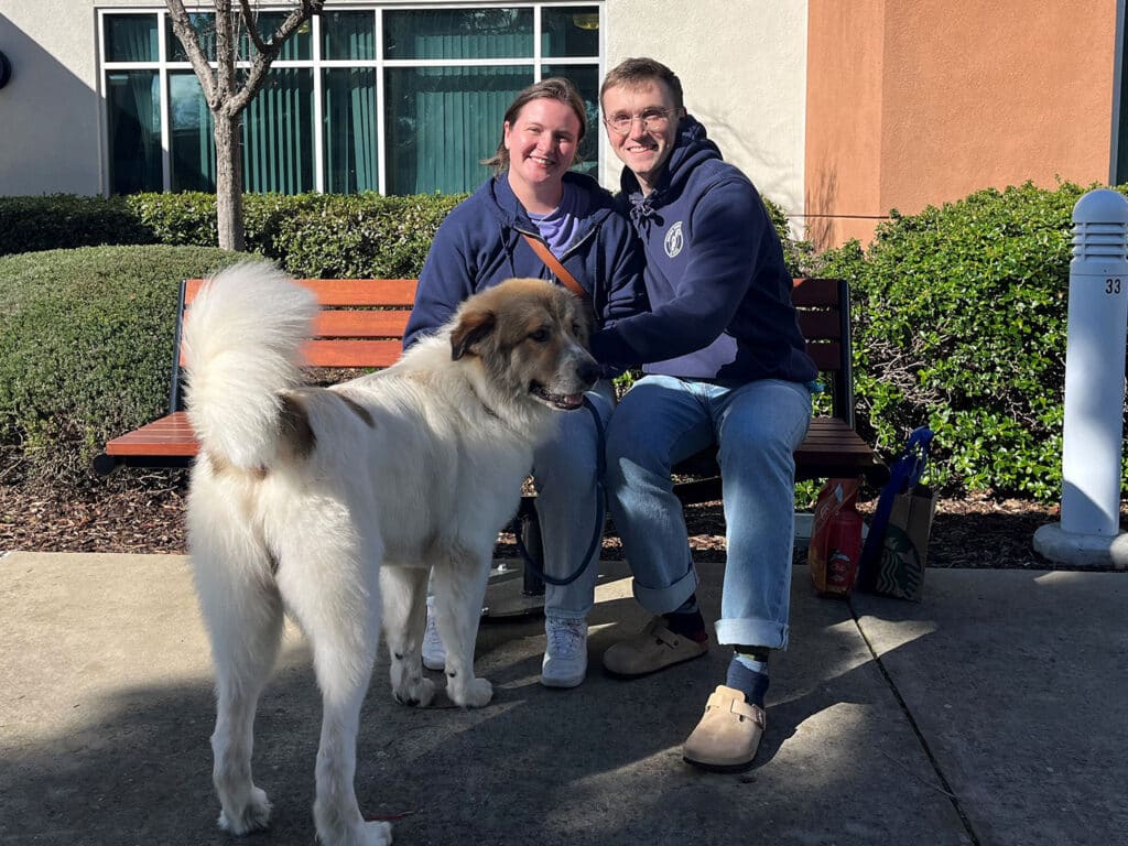 A couple sitting on a bench outdoors with a large, fluffy dog beside them. The man in a navy sweatshirt and the woman in a pink shirt are smiling. Green shrubs and buildings are visible in the background, suggesting an adoption event.