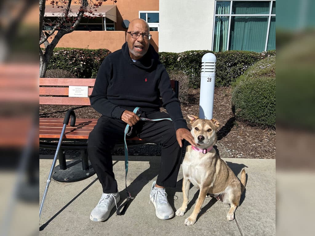 An elderly man wearing glasses sits on a wooden bench with a cane beside him. He has a friendly expression and is holding a leash attached to a light brown dog, who is sitting beside him. The setting includes greenery and a building in the background, emphasizing themes of companionship and adoption.