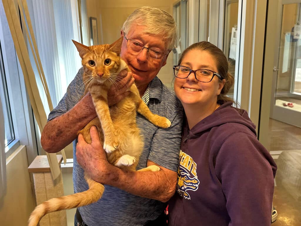 An elderly man holding an orange cat and smiling alongside a woman wearing glasses and a purple hoodie, both inside an animal shelter. This image emphasizes the joy of pet adoption.