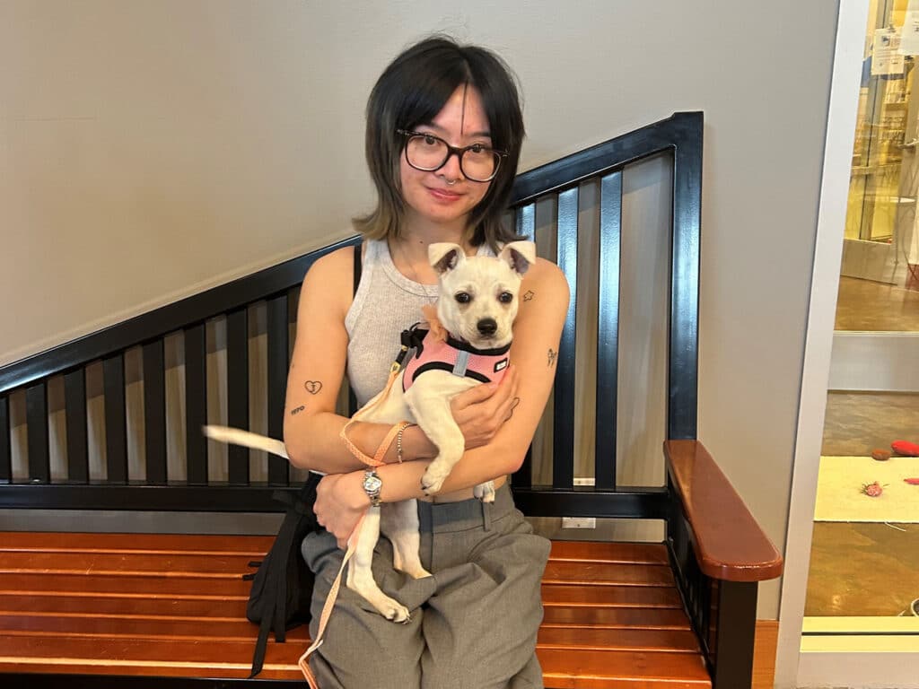 A young woman with glasses wearing a gray tank top and beige pants, holding a small white dog in her lap. They sit on a wooden bench with a modern railing behind them, in a bright indoor setting, highlighting their connection and the theme of Adoption.