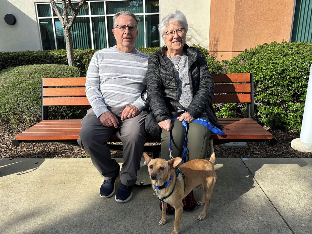 An elderly couple sitting together on a wooden bench outside a building, each wearing casual clothing. A small tan dog on a leash in front of them. The scene suggests companionship and highlights the theme of pet adoption.