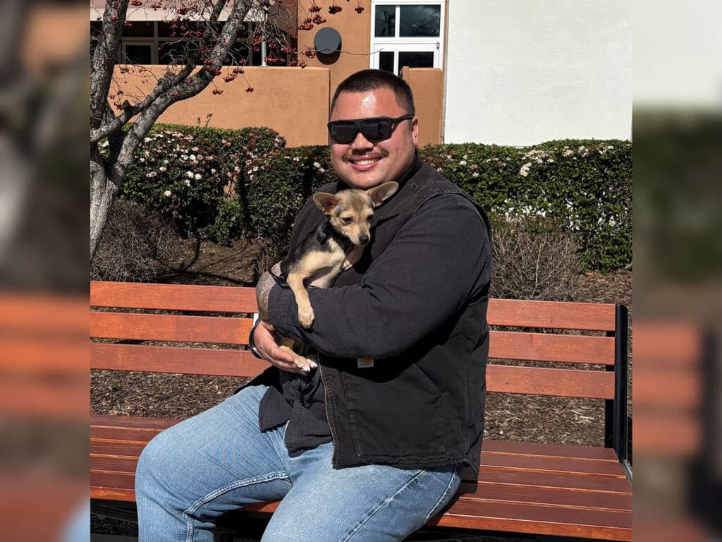 A man wearing sunglasses sits on a bench, holding a small dog in his arms. They are surrounded by greenery and a light-colored building in the background, illustrating the theme of pet adoption.
