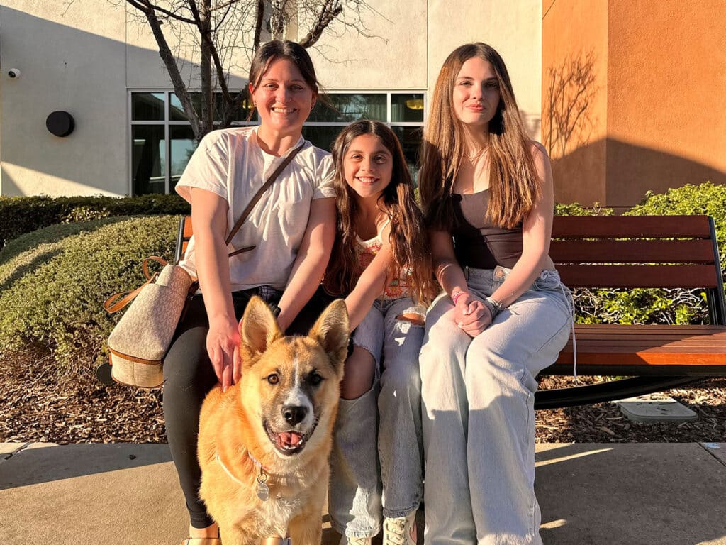 A woman and two girls sit on a bench outside a building, with a dog in the foreground. The woman smiles, wearing a light shirt and carrying a bag, while the girls, one gesturing playfully, wear casual clothing. The scene represents companionship, and highlights the theme of Adoption.