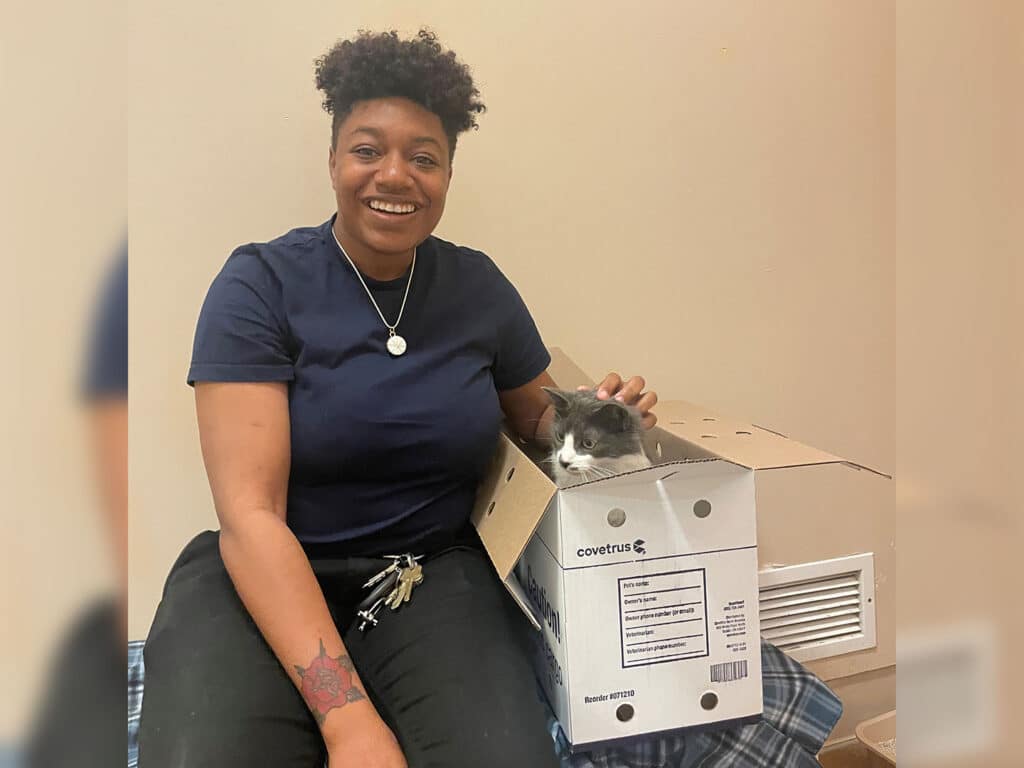 A woman with short hair and a blue shirt smiles while sitting on a bed, holding a gray and white cat that is peeking out of a box labeled "covetrus." The setting appears to be a cozy room, possibly related to animal adoption efforts.