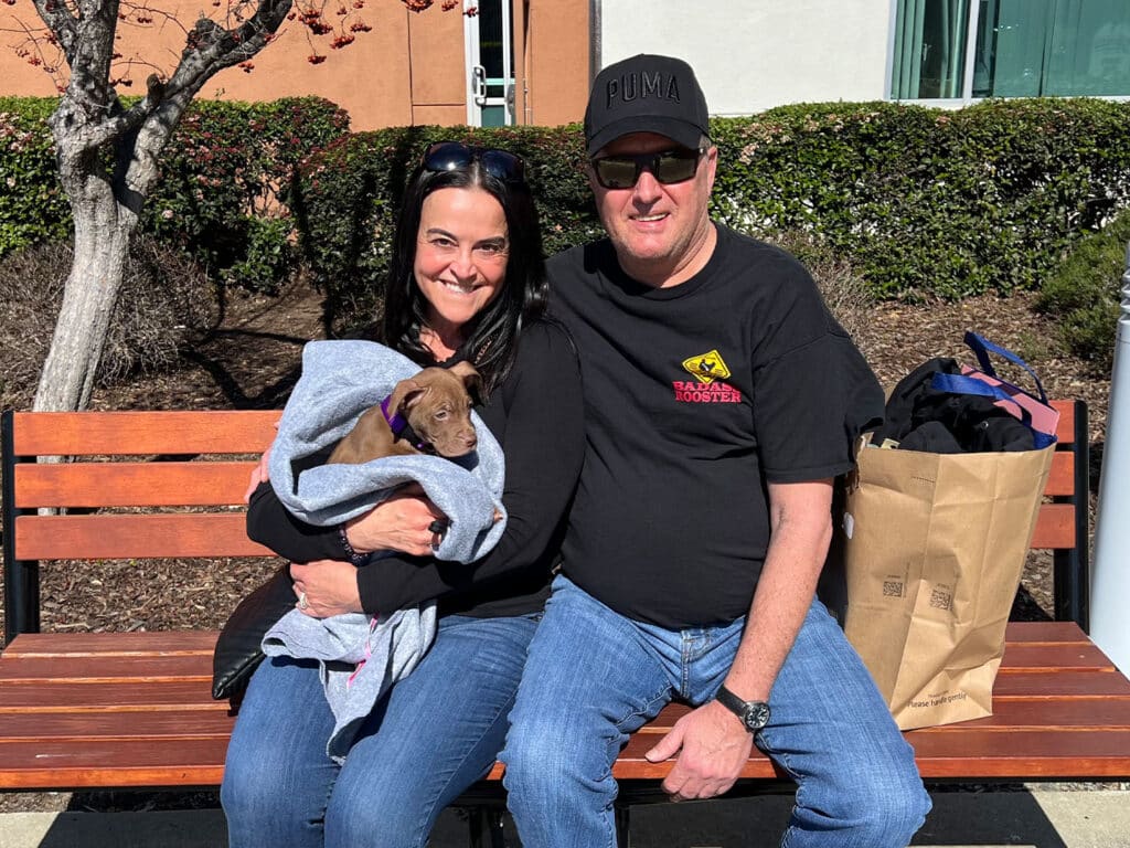 A woman and a man sit on a bench, smiling, with a small puppy wrapped in a gray blanket in the woman's arms. The man is wearing a black t-shirt and a cap. They are surrounded by greenery and sunlight, suggesting a joyful adoption moment.