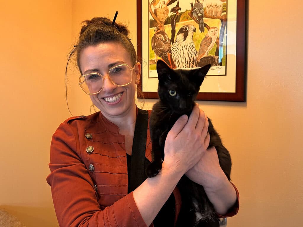 A woman wearing glasses smiles while holding a black kitten in her arms. The background features an animal-themed artwork on the wall. This image highlights the theme of Adoption.