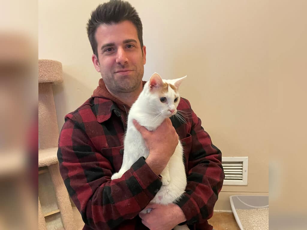 A man in a red plaid shirt holds a white cat in his arms, smiling slightly. The setting appears to be a cozy room with a cat tree and a litter box in the background. The image relates to themes of pet adoption.