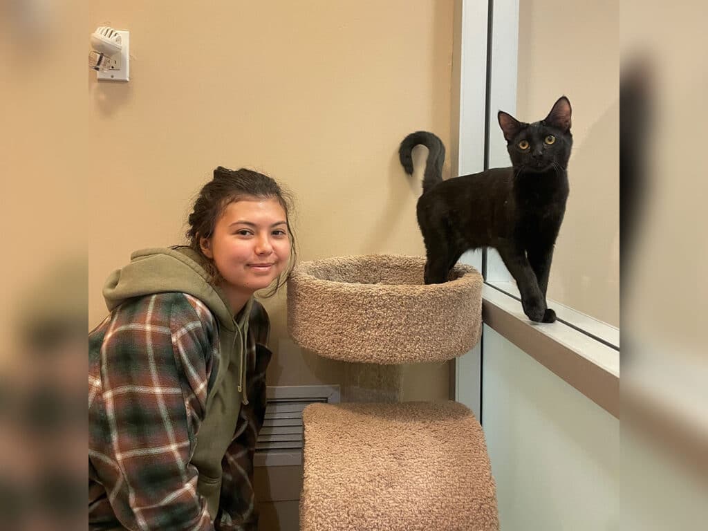 A girl with dark hair wearing a plaid jacket smiles beside a cat standing on a cat tree. The black cat looks towards the camera, while the background shows a soft beige wall and a window. This image highlights themes of adoption and companionship.
