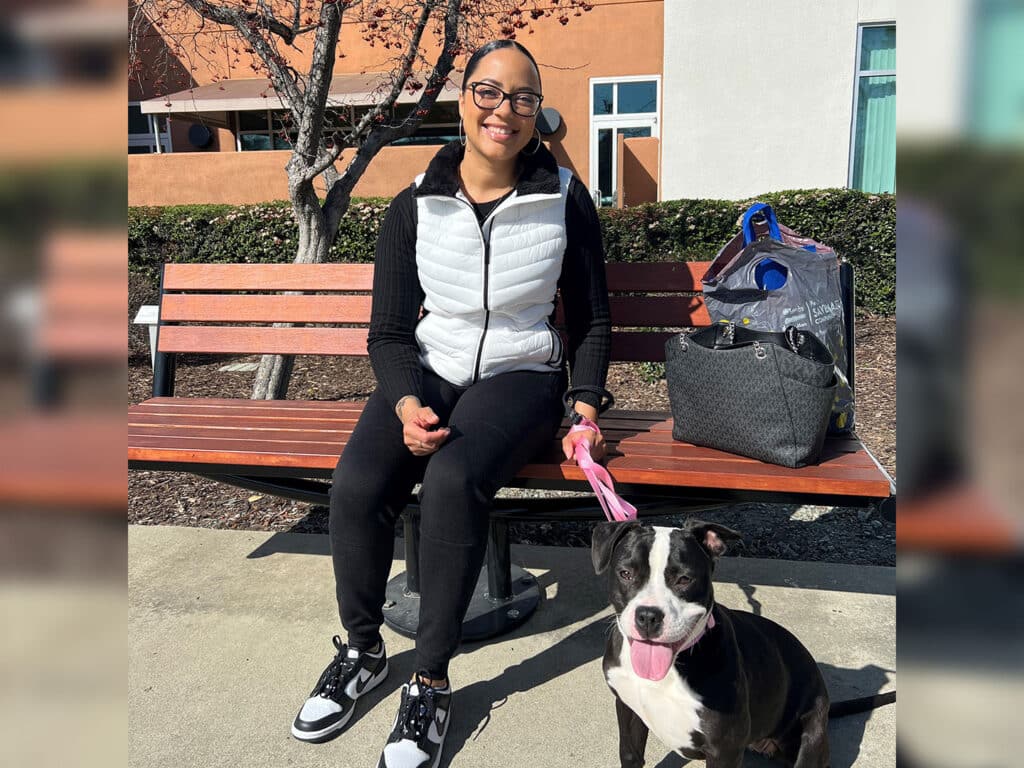A woman sitting on a bench with a black dog on a leash beside her. She is wearing a light jacket and black pants, smiling at the camera. A bag is placed next to her on the bench, surrounded by greenery, highlighting a moment of connection and adoption.