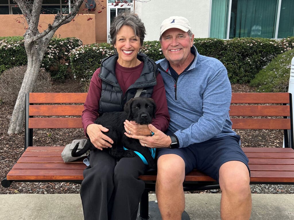 A woman and a man sitting on a wooden bench, smiling while holding a black puppy. The woman has shoulder-length hair, wearing a black vest over a burgundy shirt, while the man is wearing a cap and a blue long-sleeve shirt. The scene suggests a joyful moment of adoption.