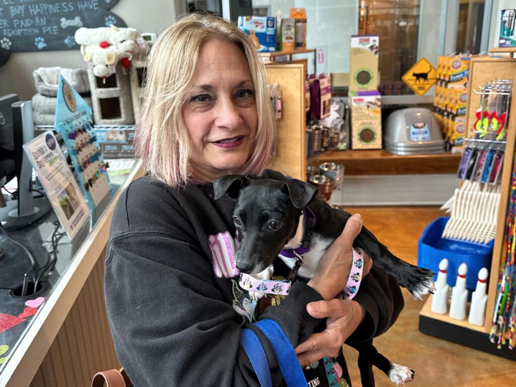 A woman with shoulder-length hair and highlighted strands holds a small black dog in her arms. The background features pet products and adoption materials in a retail setting.