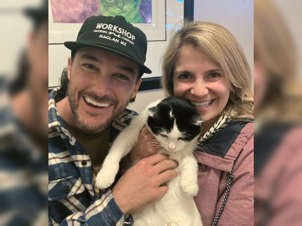 A man and a woman smile while holding a black and white cat between them. The man wears a plaid shirt and a black cap, and the woman has blonde hair and a light sweater. The background includes colorful artwork, suggesting a community space related to pet adoption.