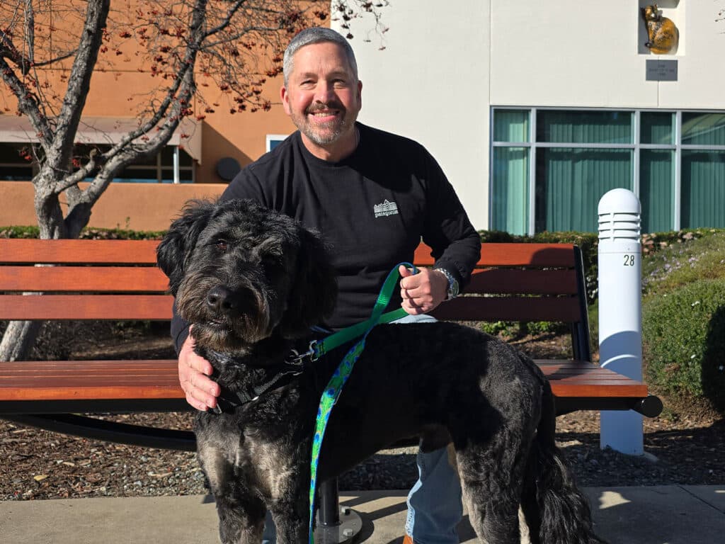 A man sitting on a bench smiles while holding a large black dog on a leash outside a building. The setting includes trees and a concrete pathway, suggesting a community environment. The image emphasizes the theme of adoption, showcasing the bond between the man and the dog.