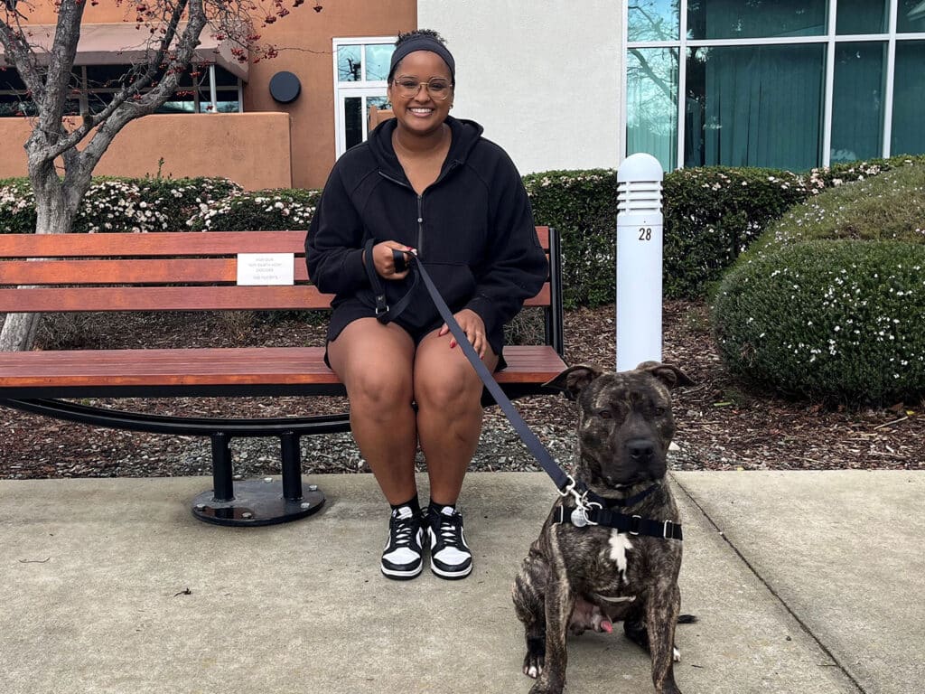 A woman sitting on a bench beside a brown and black striped dog on a leash. Both are looking at the camera. The background features trees and a building, suggesting an outdoor adoption event.