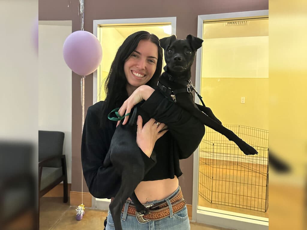 A smiling woman holds a small black dog in her arms inside a cozy room with light-colored walls. The background features a glass-paneled area likely for pet interactions, and a pink balloon is visible in the corner. Adoption is a potential theme.