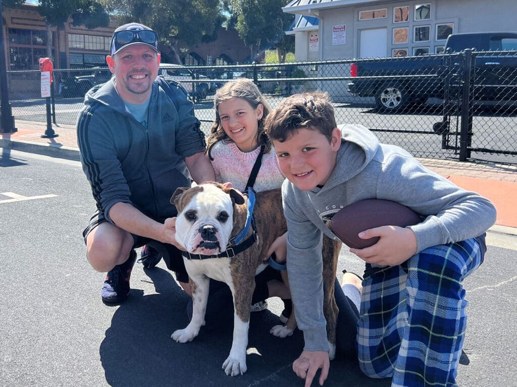 A man, a girl, and a boy pose with a brown and white dog on a sunny day. The man and children are smiling, and the girl is petting the dog. The boy holds a football, and they are in a parking area near buildings, suggesting community engagement or adoption event.