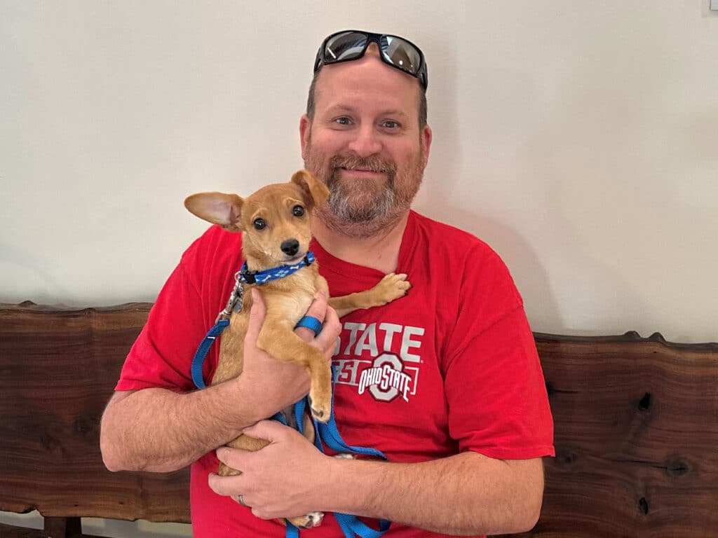 A man with a beard holding a small tan dog with a collar, sitting on a wooden bench. The man is wearing a red T-shirt and sunglasses. The background is a light-colored wall, and the dog appears to be calm and comfortable in his arms, symbolizing the theme of adoption.
