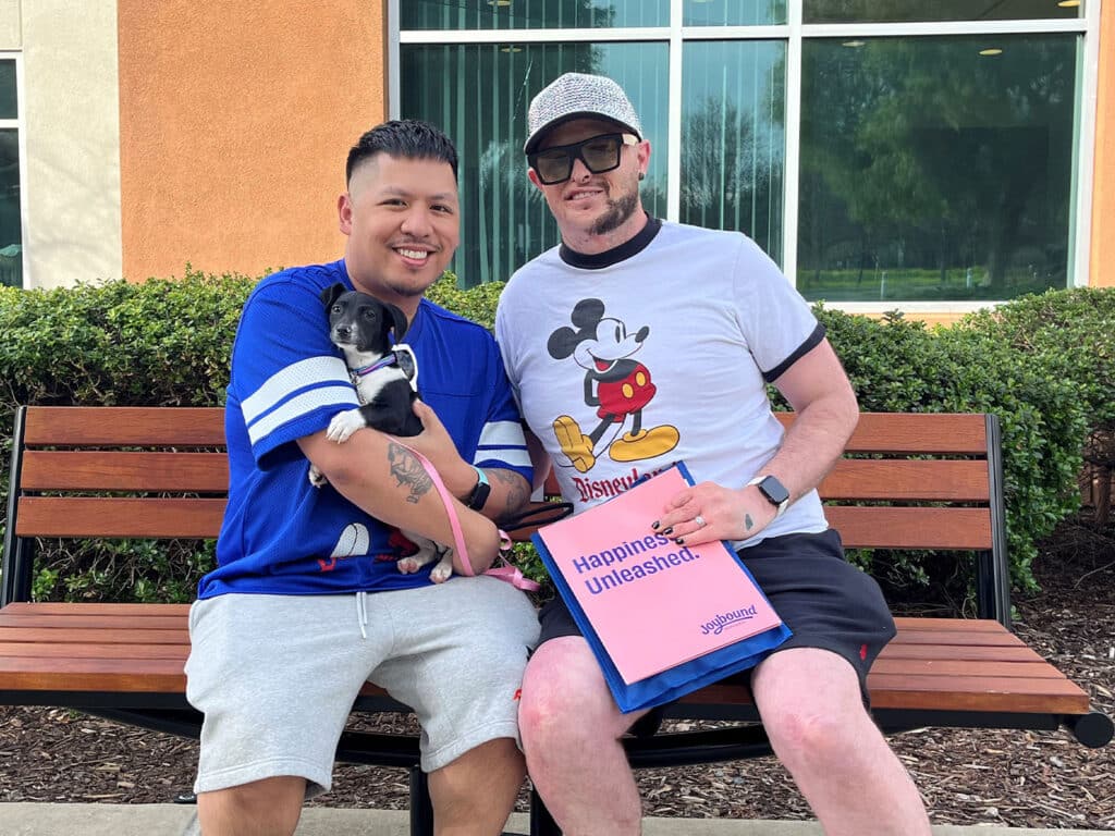 Two men sitting on a wooden bench outside, one holding a small black and white puppy. The other man is wearing a Mickey Mouse T-shirt and holding a sign that reads 'Happiness Unleashed.' They are in a park-like setting, surrounded by greenery.