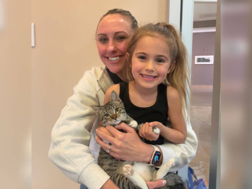 A woman smiling while holding a young girl and a gray tabby cat. The girl has long hair and is dressed in a black tank top, resting the cat on her lap. The scene takes place indoors, with light-colored walls and a door visible in the background. The image relates to adoption.