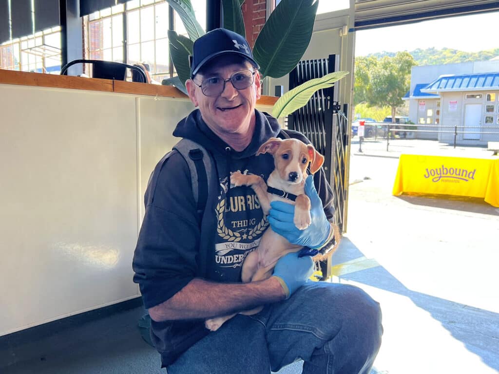 A man wearing glasses and a black cap is holding a small puppy in a blue sweater inside a brightly lit room. Behind them, there is a table with the Joybound logo visible, and a green plant is positioned nearby.