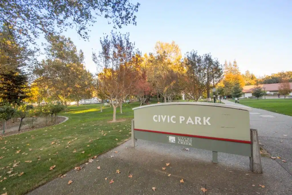 A sign reading "Civic Park" in a landscaped area with trees displaying autumn foliage. The park features grassy lawns and winding pathways, indicating a tranquil outdoor setting.