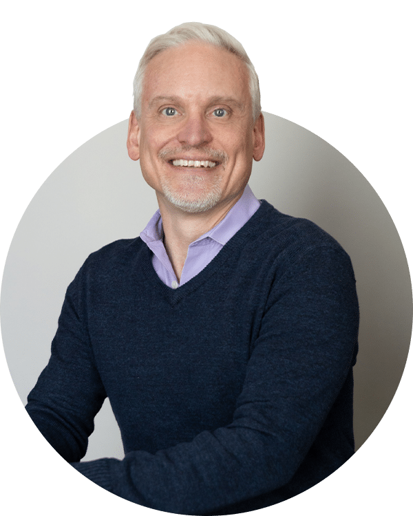 A smiling man with light gray hair wearing a dark blue sweater over a light purple shirt, seated at a table. The background is neutral in color. This image represents a member of the Joybound Team.