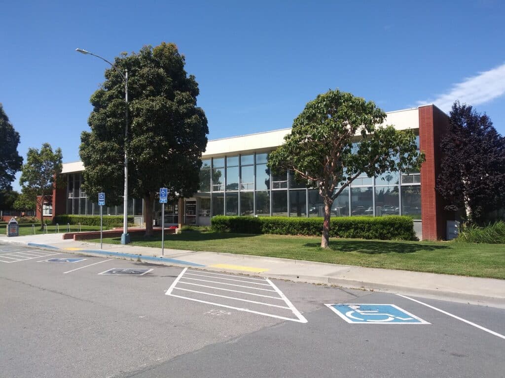 A modern building with large windows, surrounded by green shrubs and trees. The parking lot features several accessible parking spaces. The scene is set under a clear blue sky, contributing to a welcoming atmosphere for pet vaccinations.