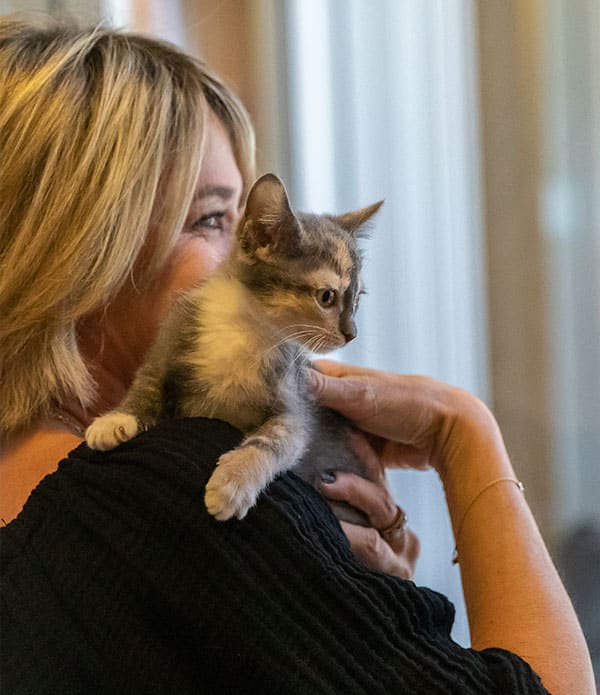 A woman with shoulder-length blonde hair holds a small gray and white kitten against her shoulder, looking at the kitten with focus. The background is softly blurred, suggesting an indoor setting.