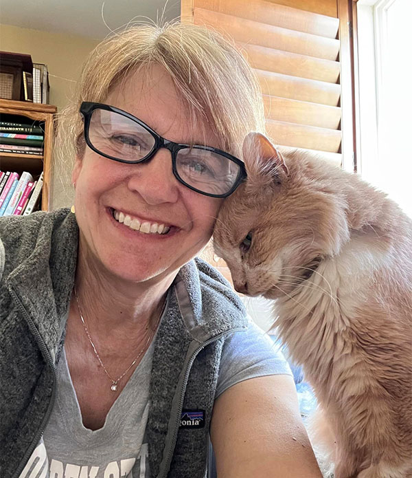 A smiling woman with glasses leans her head against an orange cat in a cozy indoor setting. They appear content and connected, highlighting themes of companionship and fostering relationships with pets.