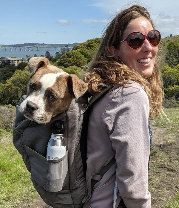 A woman wearing sunglasses smiles while hiking, carrying a dog in a backpack designed for pets. The dog's head peeks out, and a water bottle is secured to the side of the backpack. A scenic view of trees and a body of water is visible in the background.
