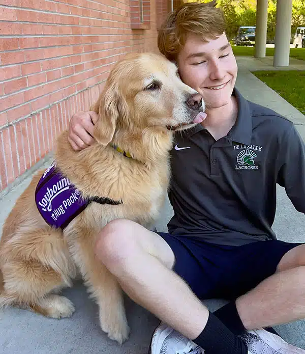 A young man sitting on the ground with a golden retriever wearing a purple vest labeled "Comfort Dog Team". The man smiles while petting the dog, with a brick building and greenery in the background. The scene reflects a volunteer activity involving therapy animals.