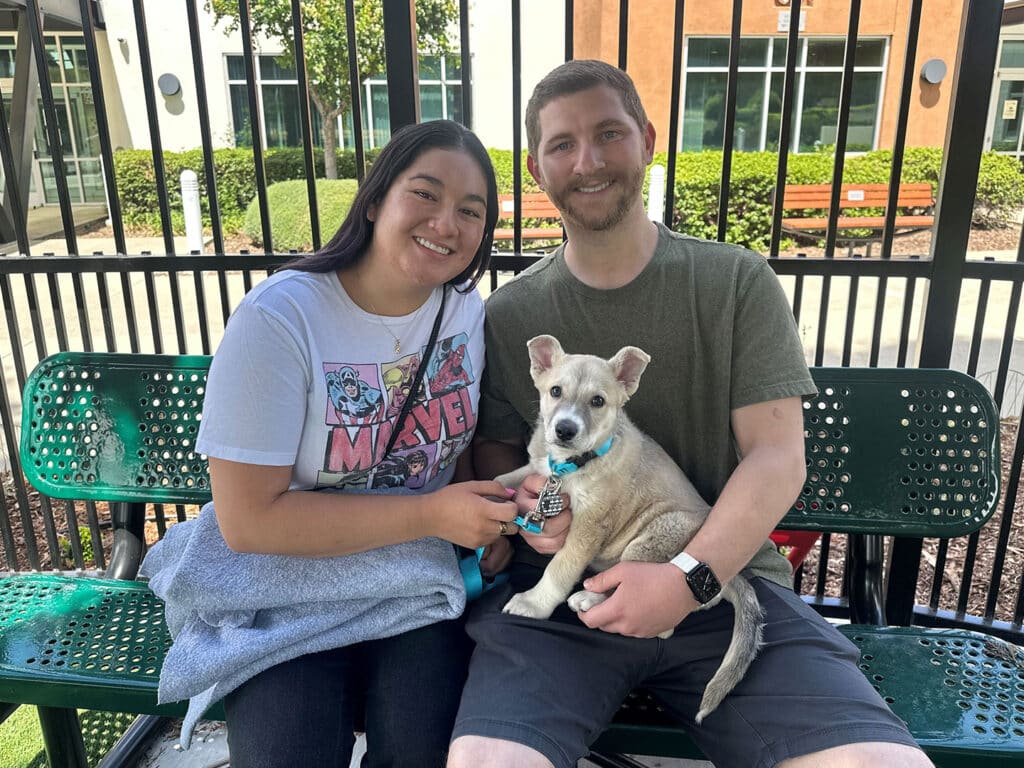 A couple sitting on a green bench in a park, smiling while holding a small light-colored dog on their laps. The woman wears a Marvel graphic t-shirt, and the man is in a gray shirt. Their adoption of the dog is evident in their joyful expressions.