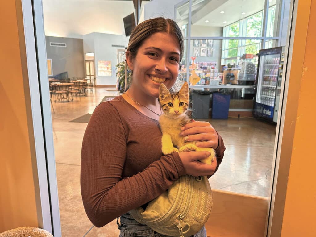 A young woman wearing a brown long-sleeve shirt holds a small kitten in a bag. They are standing indoors in a spacious area with large windows and seating in the background, highlighting a moment of adoption.