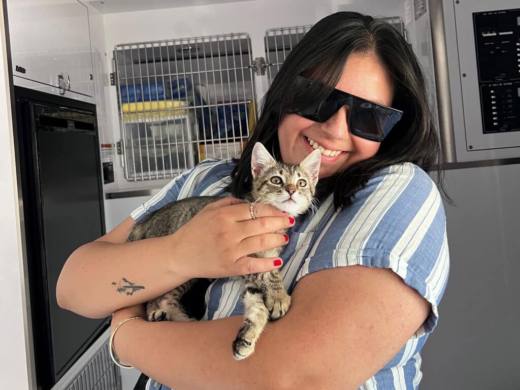 A woman wearing sunglasses smiles while holding a small gray tabby kitten, showcasing a joyful moment. The background includes enclosures, suggesting a pet adoption setting, emphasizing the theme of adoption.