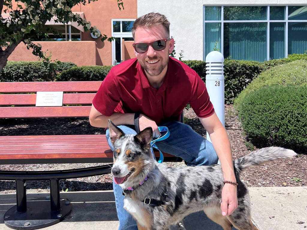 A man wearing sunglasses kneels beside a dog with a speckled coat in an outdoor area with a bench. The setting appears bright and sunny, suggesting a recent adoption. The man smiles while holding the dog's leash.